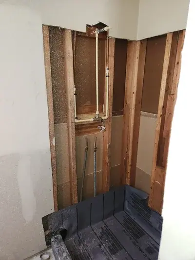 A bathroom under renovation shows exposed wooden wall studs with copper and PEX plumbing pipes installed for a shower fixture. A black shower pan liner is visible at the bottom, and drywall is partially installed on the left wall.