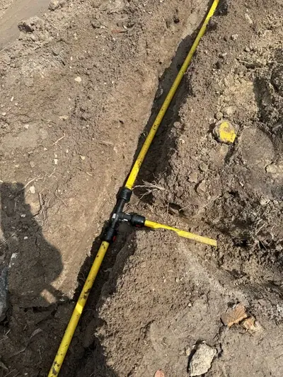 A yellow utility pipe with a black T-connector is laid in a dirt trench. A branch pipe extends from the connector into the disturbed ground.