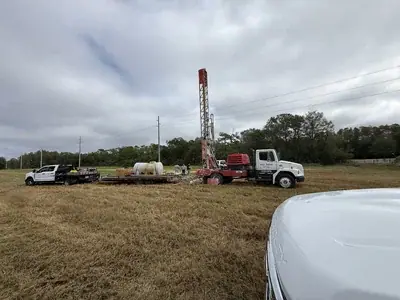 A large red and white drilling rig truck is parked in a dry grassy field, with a white pickup truck and trailer nearby. Power lines, utility poles, and trees line the background under a cloudy sky, with a few people visible near the drilling equipment.