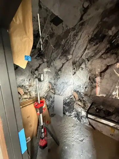 An unfinished room with walls covered in black and white marble-patterned material, featuring a work light on a tripod and cardboard boxes on the floor. A glass panel reflects a person and a ladder, while a dark counter sits on the right.