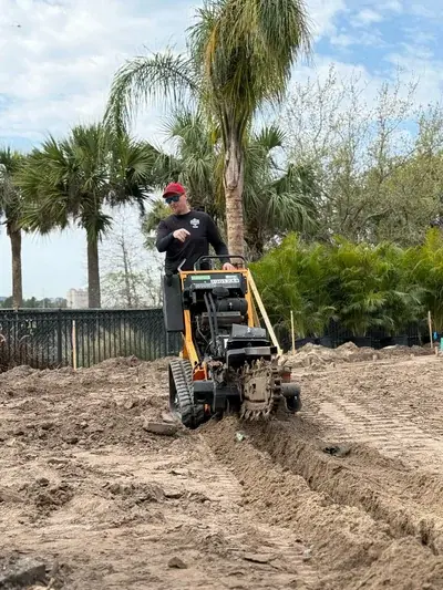 A man in a red hat and sunglasses operates a yellow trencher machine, digging a trench in a dirt field. Palm trees and other foliage are visible in the background under a cloudy sky.