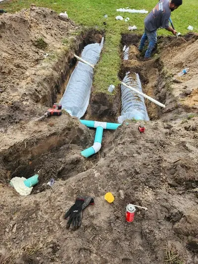 A construction site shows two long trenches dug in the dirt, with corrugated pipes wrapped in white fabric laid inside them. A worker is shoveling dirt in the background, while various plumbing pipes and tools are scattered around the trenches.