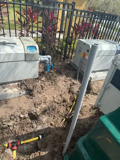 An outdoor photo showing two large grey utility units, one labeled 'Lochinvar', with disturbed earth and exposed piping around their bases. Grey PVC pipes with blue valves and yellow flexible piping are visible, set against a black metal fence and red-leafed plants.