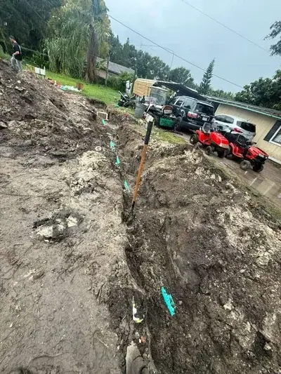 A muddy construction site features a long trench with blue pipes and a shovel stuck in the dirt. In the background, a person walks on a dirt mound, and several vehicles, including two red riding lawnmowers, are parked near a house.