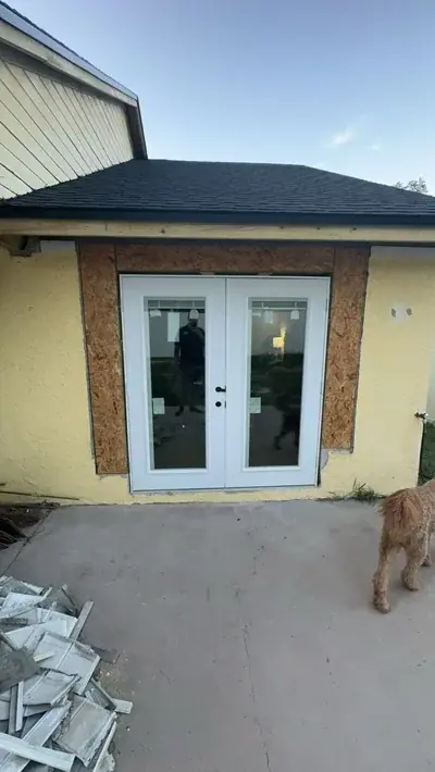 A newly installed white double door is set into a yellow stucco wall with exposed OSB sheathing around the frame. A new black shingle roof is visible above, and construction debris is piled on the concrete patio to the left.