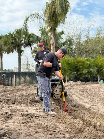 Two men are working outdoors on a construction site. One man in the foreground is digging a trench with a red and yellow shovel, while another operates a small tracked machine in the background, surrounded by dirt and palm trees.