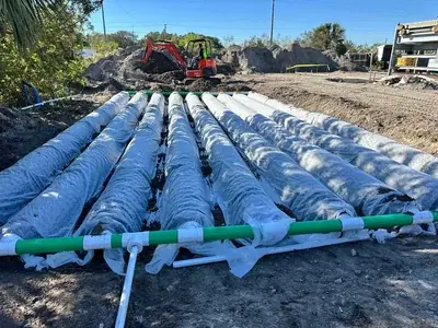 Multiple large, white-wrapped pipes are laid in a trench at a construction site, with a green and white pipe assembly visible in the foreground. An excavator is actively digging in the background, surrounded by piles of dirt.