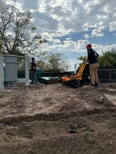 Two men are working on a construction site under a cloudy sky. One man operates an orange stump grinder on a mound of dirt, while another stands near green structures and metal panels in the background.