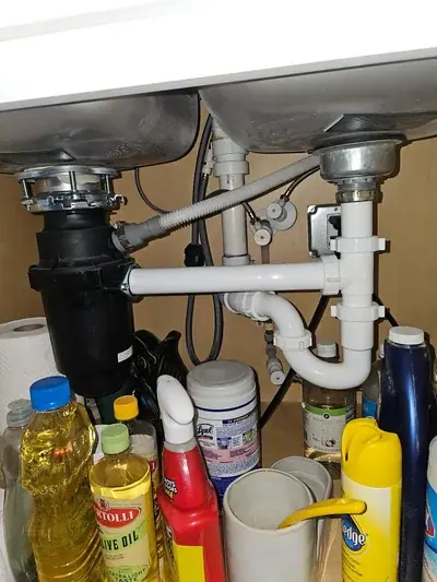 A view under a kitchen sink showing the plumbing for a double basin, including a black garbage disposal unit on the left and white PVC pipes connecting both drains. Various cleaning supplies and bottles are visible on the cabinet floor.