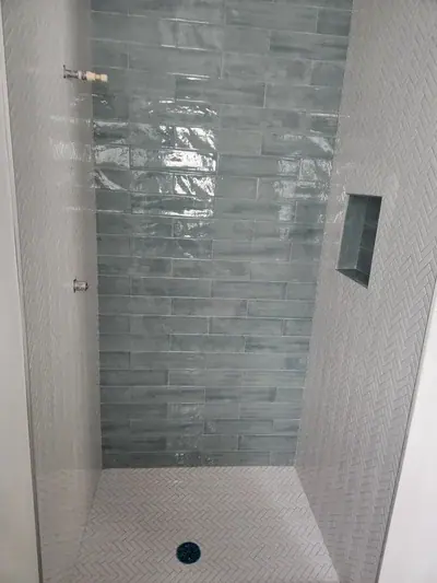 A newly tiled shower stall features a back wall with glossy, light blue-green rectangular tiles. The side walls and floor are covered in light gray herringbone-patterned tiles, with a recessed niche on the right and pipe stubs on the left.