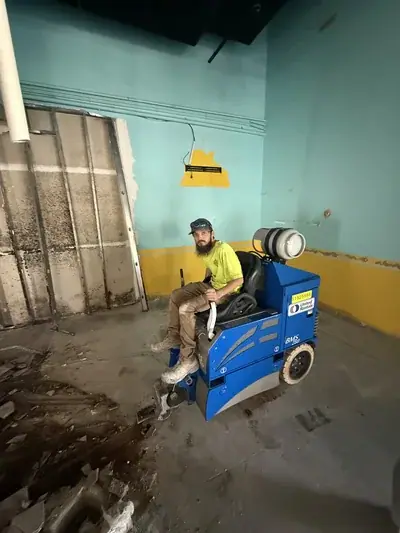 A man with a beard and baseball cap sits on a blue ride-on floor scraper machine, identified as a United Rentals BMS 280, in a room under renovation. The room features light blue and yellow walls, exposed metal wall framing, and a concrete floor with debris.