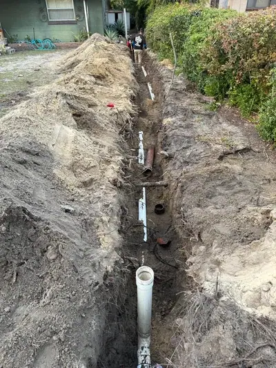A long, deep trench is dug in the ground, with piles of dirt on either side. White PVC pipes and older brown pipes are visible along the bottom of the trench. A worker is seen in the background walking along the trench.