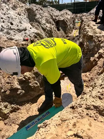A plumber in a hard hat and a yellow shirt with a company logo is bent over, working on a green pipe in a dirt trench. A level tool rests on the pipe, indicating work in progress.