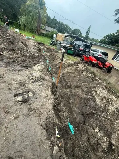 A long, muddy trench with light blue pipes and a shovel stuck upright in the center. In the background, a person walks up a dirt slope, and several vehicles including two red riding lawnmowers are parked near a house.