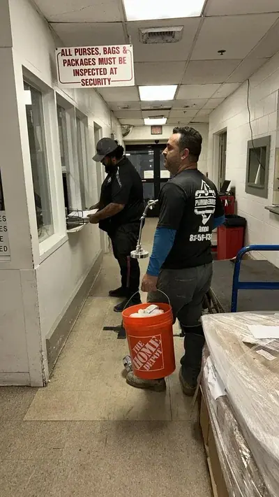 Two men are in a hallway, one standing at a counter with papers and the other holding an orange Home Depot bucket and a chrome fixture. A sign above the counter reads "ALL PURSES, BAGS & PACKAGES MUST BE INSPECTED AT SECURITY".
