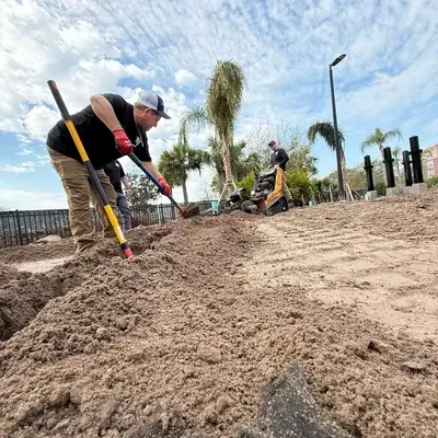 Two workers are digging a trench in sandy soil under a partly cloudy sky. One worker uses a shovel in the foreground, while another operates a small trenching machine in the background, with palm trees and a street light visible.