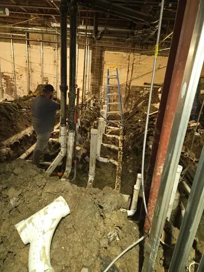 A plumber in a dark shirt and cap is working on a network of white PVC and black cast iron pipes in a muddy trench. Overhead, more pipes are visible, and a blue ladder leans against a wall in the background.