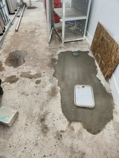 A concrete floor with several wet spots and a fresh patch of darker gray concrete containing a round green cover and a rectangular white utility cover. Industrial pipes, a metal mesh cage, and a piece of plywood are visible in the background.