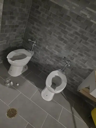 Two white toilets with open seats are positioned on a light gray tiled floor against dark gray subway tile walls. Scattered white plastic caps are visible on the floor near the left toilet, and a floor drain is in the bottom left.