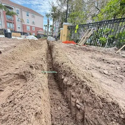 A long, narrow trench is dug into sandy soil, with a green pipe visible crossing the trench. In the background, apartment buildings and a black metal fence are visible under a partly cloudy sky.