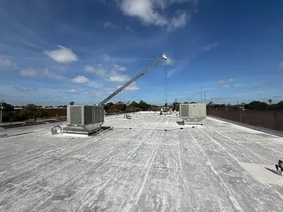 A wide shot of a flat commercial rooftop under a blue sky with scattered clouds. Several HVAC units are visible, along with a crane extending over the roof and workers in the distance.
