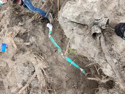 A person's legs and feet are visible in a muddy trench with exposed plumbing pipes. A turquoise pipe with a white fitting connects to older, darker pipes in the dirt, surrounded by soil and roots.