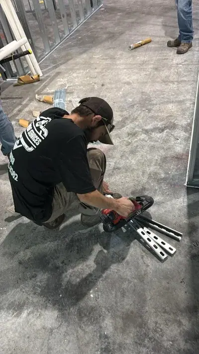 A worker wearing a black t-shirt and a baseball cap crouches on a concrete floor, using a red power tool to cut metal channels. Metal framing studs are visible in the background of the construction site.