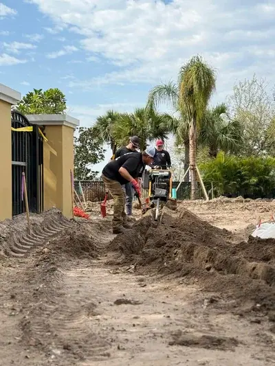 Three men are working outdoors, with one operating a trenching machine to dig a trench in the dirt. Piles of dirt line the newly dug trench, with a building and palm trees visible in the background under a cloudy sky.