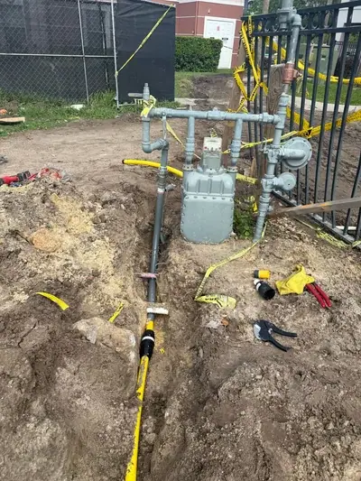 A gas meter and associated piping are visible next to a black metal fence, with a yellow gas line extending into an open dirt trench. Yellow caution tape is wrapped around the fence and pipes, and various tools are scattered on the ground.