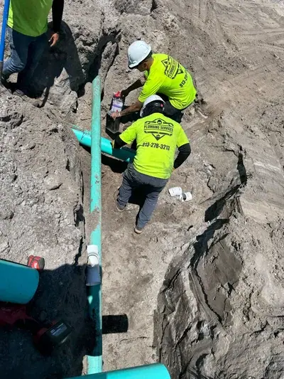 Two plumbers in hard hats and neon yellow shirts are working in a dirt trench, installing or repairing turquoise pipes. One worker is applying a substance from a purple container to a pipe section.