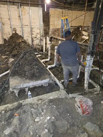 A plumber stands in a muddy, excavated area, surrounded by newly installed white PVC pipes and existing black pipes. Piles of dirt and concrete debris are visible, with a blue ladder in the background.