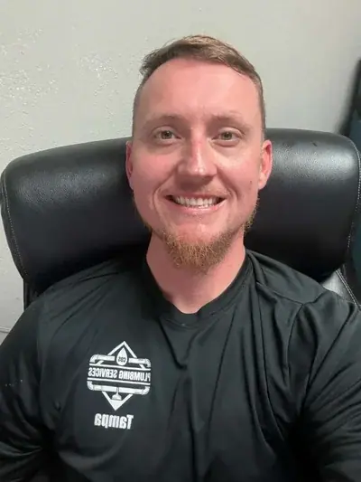 A smiling man with short brown hair and a reddish-brown goatee sits in a black office chair. He wears a black long-sleeved shirt with a mirrored white logo that reads 'PLUMBING SERVICES' and 'Tampa'.