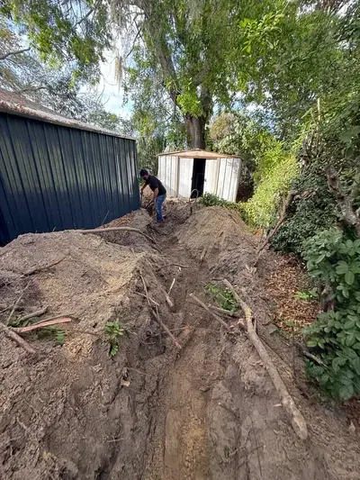 A worker in a black shirt and jeans is digging a trench in a dirt path between a dark green metal shed and a smaller white metal shed, surrounded by trees and foliage.