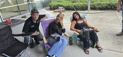 Three people are gathered outdoors on a concrete surface. A man crouches on the left, while two women sit in beach chairs, all smiling towards the camera, with construction equipment visible in the background.
