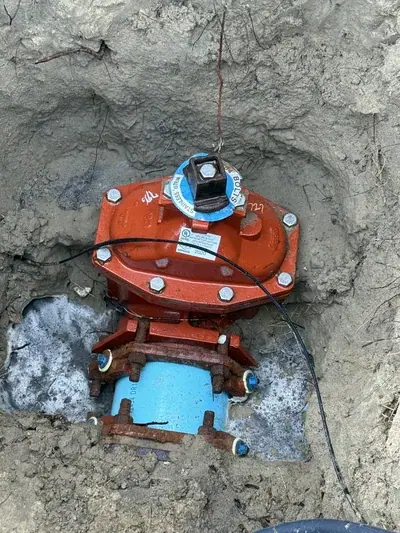 A partially buried orange-red fire hydrant valve assembly is shown in a pit of light-colored soil. It features a blue pipe section at the bottom, connected by rusty bolts, with a black wire and a thin copper wire attached to the top of the valve.
