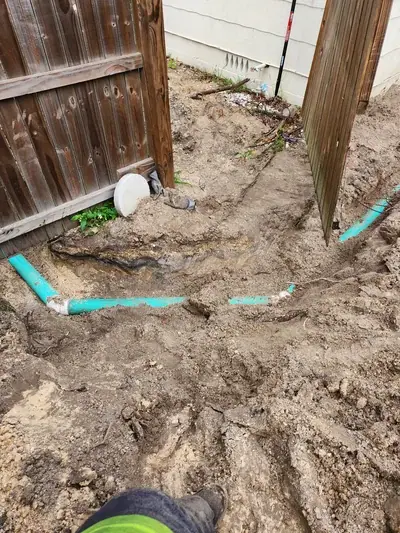 A trench dug in muddy dirt reveals light blue pipes laid underground, with a wooden fence on either side and a house wall in the background.