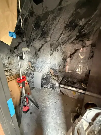 A room under construction with walls covered in dark and light patterned stone, featuring a work light on a tripod, cardboard boxes, and a glass shelf with various items.