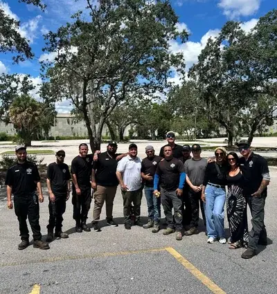 A group of 14 people, mostly men, stand in a paved outdoor area under a blue sky with white clouds. Large trees with Spanish moss are visible in the background.