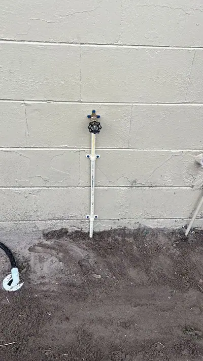 A white PVC pipe extends vertically from the dark brown soil, secured to a light-colored block wall with two blue clips, and topped with a spigot featuring a black handle and blue cap. A black hose with a white connector lies on the ground to the left.