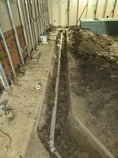 A long trench is dug into the dirt floor of an indoor construction site, revealing white PVC plumbing pipes laid within. Metal wall studs line the left side, and a stack of drywall sheets is visible in the background.