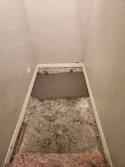 A narrow hallway or landing with light-colored walls and white baseboards. The floor is partially covered with a section of wet, dark gray concrete or leveling compound, while the front half shows a dirty, exposed subfloor with debris and a small section of carpet padding at the bottom.
