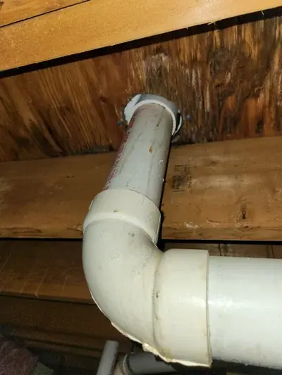 A white PVC pipe system is shown, with a vertical section passing through a wooden board and connecting to a horizontal section via an elbow joint. The surrounding wooden surfaces and the pipe itself exhibit signs of water staining and discoloration.