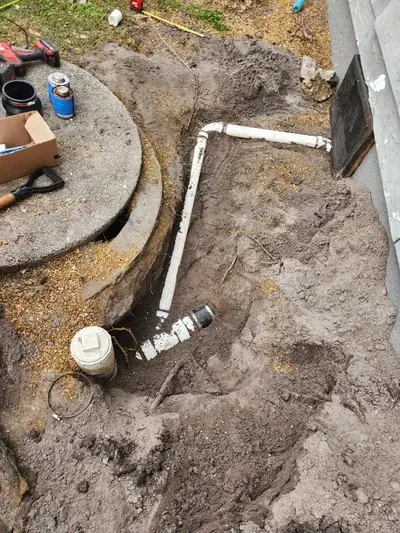 An excavated trench in dirt shows white PVC pipes being installed or repaired, with a concrete manhole cover and plumbing tools nearby.