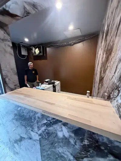 A man in a t-shirt and cap stands behind a light wooden countertop in a room with marble-patterned walls and a plain brown wall. Exposed wires and network equipment are visible on the upper wall and ceiling, suggesting a space under construction.