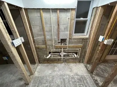 The image displays exposed plumbing pipes, including white PVC and beige PEX, installed against a concrete block wall. Wooden studs frame the area, and a window is visible on the right side, indicating a renovation or construction stage.