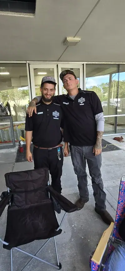 Two men in black polo shirts with company logos stand outside a building with glass doors. The man on the left, with a beard and grey cap, has his arm around the shoulder of the man on the right, who wears a camouflage cap and has tattoos.