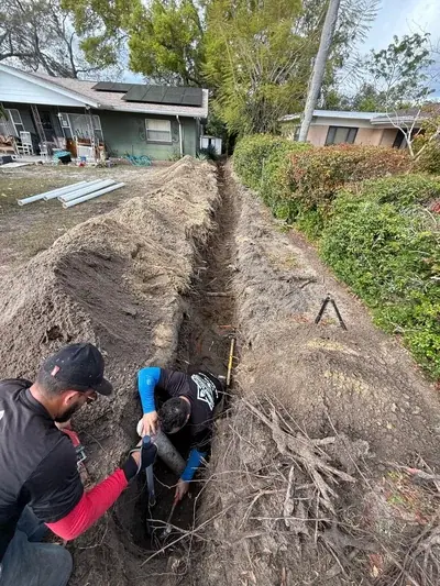 Two plumbers are working in a long, deep trench in a residential yard. One plumber is in the trench connecting a pipe, while the other assists from above, with piles of excavated dirt on either side.