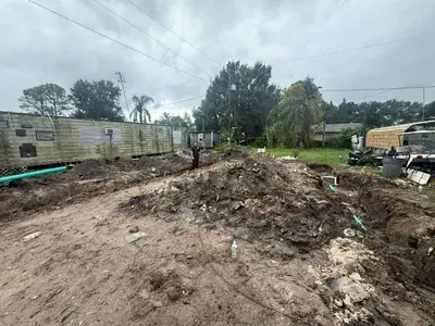 A muddy construction site with trenches and dirt piles in the foreground, showing green and white pipes. In the background, a weathered mobile home is on the left, and a partially disassembled RV or trailer is on the right, with two men standing near the trenches under an overcast sky.