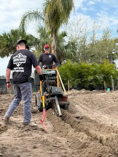 Two workers are shown in a dirt field, one operating a trenching machine to dig a trench, and another worker walking beside it. Palm trees are visible in the background under a partly cloudy sky.