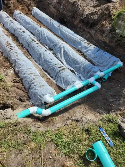 An excavated trench reveals four long, white fabric-wrapped drainage pipes partially covered with dirt. Bright turquoise PVC pipes with white fittings are laid in the foreground, connecting to the wrapped pipes, with a spirit level and pipe sections on the grassy ground.